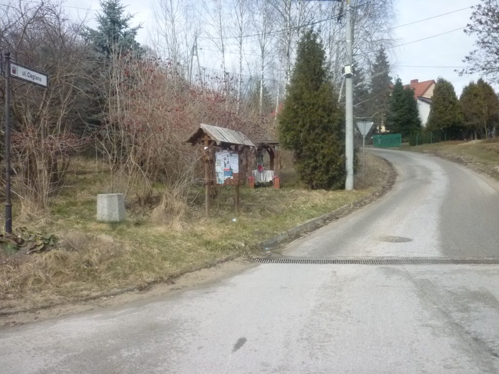 Ul. Ceglana od strony ul. Wielkowiejskiej - 2023 r. Widok na fragment ulicy asfaltowej prowadzącej w górę, po lewej tablica ogłoszeń z drzewami w tle.