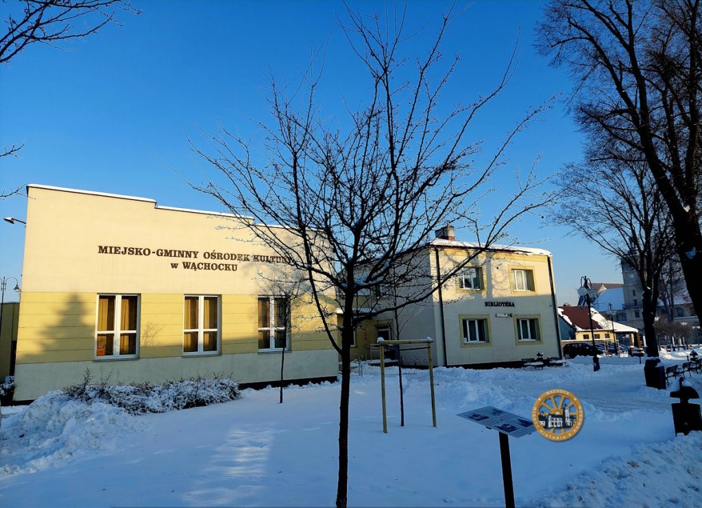 Pokryta śniegiem scena przedstawiająca Miejski-Gminny Ośrodek Kultury w Wąchocku, z czystym błękitnym niebem i nagimi drzewami. MGOK