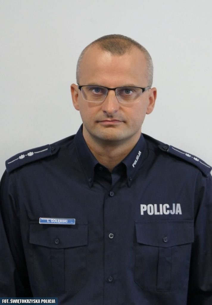 Na zdjęciu znajduje się funkcjonariusz policji w granatowym mundurze z napisem "POLICJA" na piersi. Mężczyzna nosi okulary i ma krótkie, ciemne włosy. Na mundurze widoczna jest plakietka z nazwiskiem "E. Gołębski". Funkcjonariusz stoi na neutralnym tle i patrzy prosto w obiektyw.