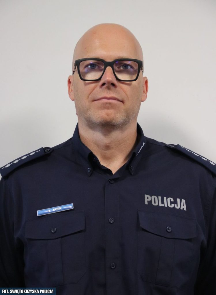 Na zdjęciu widoczny jest funkcjonariusz policji w granatowym mundurze z napisem "POLICJA" na piersi. Mężczyzna ma okulary, jest łysy i patrzy prosto w obiektyw. Na mundurze widoczna jest plakietka z nazwiskiem "E. Jasik". Funkcjonariusz stoi na jasnym, neutralnym tle.