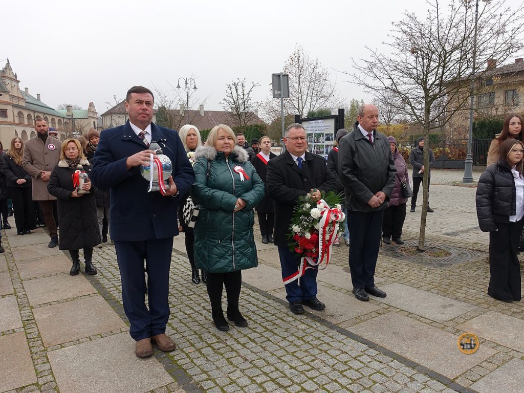 Na zdjęciu znajdują się uczestnicy obchodów święta niepodległości w Wąchocku. W pierwszym rzędzie ze zniczem oraz wieńcami stoją przedstawiciele samorządu wąchockiego, burmistrz, jego zastępca, radny oraz przewodnicząca rady miejskiej. Pogoda pochmurna, zimno, bez słońca.