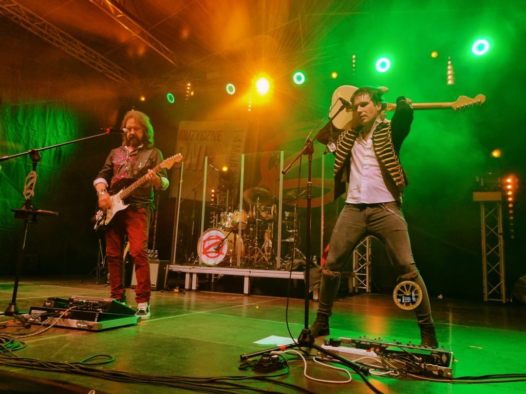 artyści na scenie, jeden gra na gitarze, drugi trzyma ją za głową, kolorowe światła dookoła