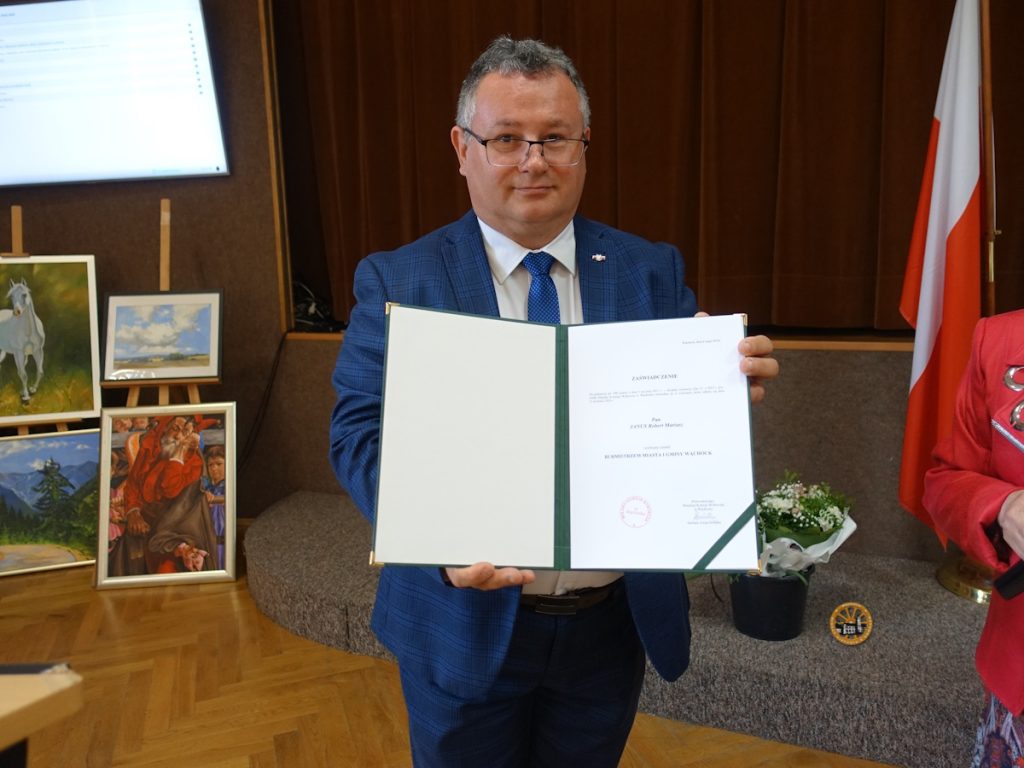 Mężczyzna w garniturze prezentuje dyplom z zaświadczeniem o objęciu stanowiska burmistrza. W tle brązowa, sceniczna kotara, flaga Polski oraz kilka obrazów.