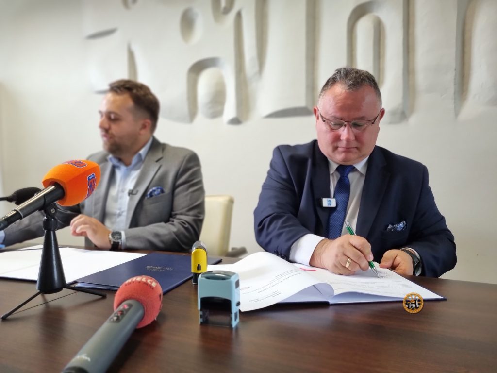 Burmistrz Miasta i Gminy Wąchock Robert Janus podpisuje porozumienie pomiędzy gminami, przed nim znajdują się mikrofony, obok siedzi mężczyzna (prezydent miasta Starachowice), przed mężczyznami leżą dokumenty