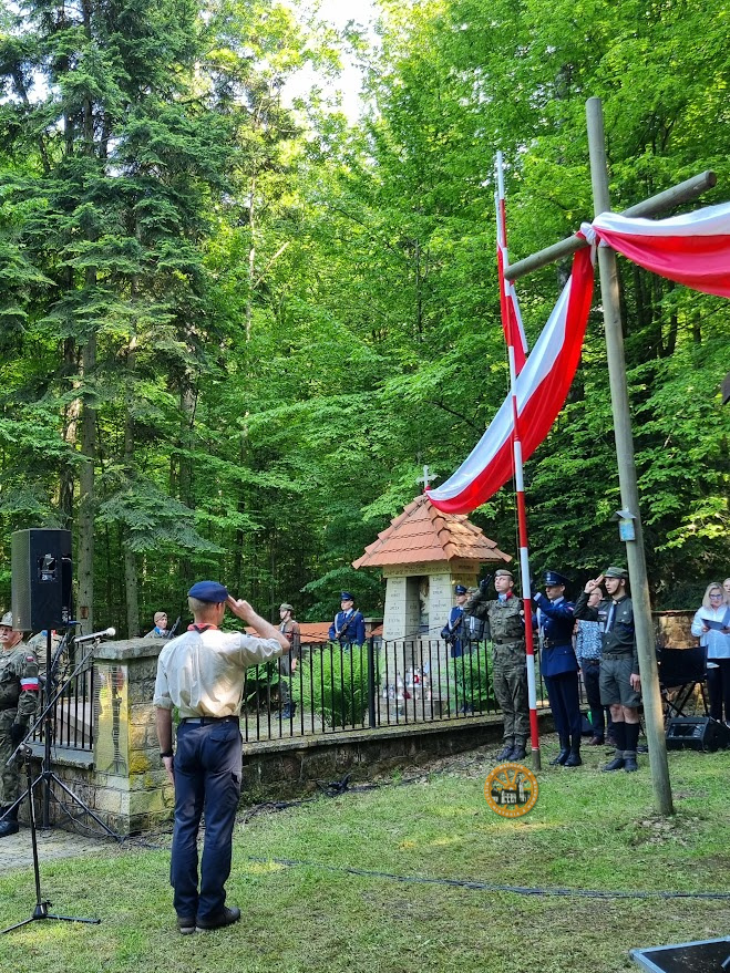 Zdjęcie przedstawia polanę wykusową z salutującymi żołnierzami i harcerzami, z flagą wciąganą na maszt