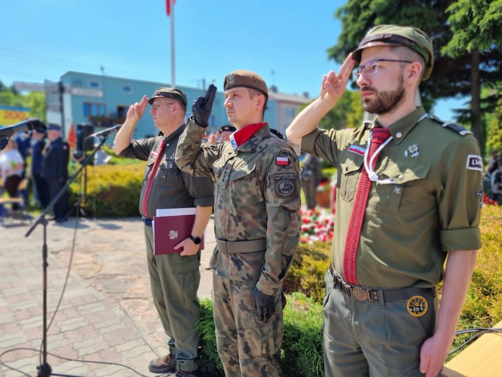 Zdjęcie przedstawia trzech salutujących mężczyzn, stoją bokiem do obiektywy, ubrani są w mundury.