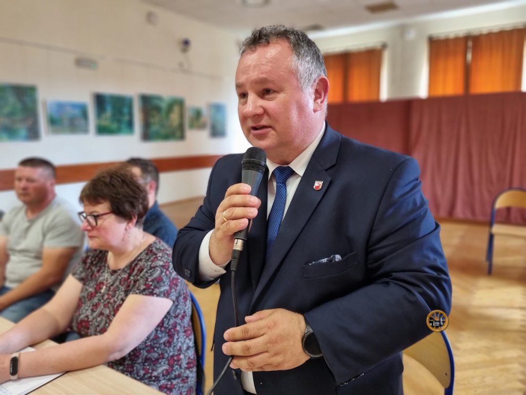 Zdjęcie przedstawia mężczyznę w garniturze, który przemawia do mikrofony, który trzyma w ręku. Obok siedzą osoby uczestniczące w sesji Rady Miejskiej