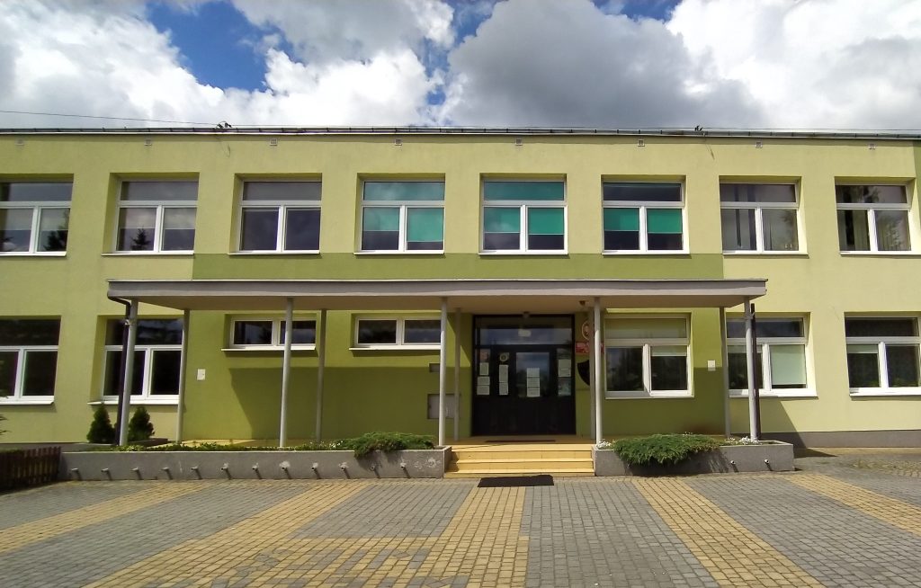 Zdjęcie przedstawia wejście do szkoły w wielkiej wsi, szkoła ma parter i piętro, do wejścia prowadzą trzy schodki, budynek w kolorze zieleni, przed nim plac z kostki