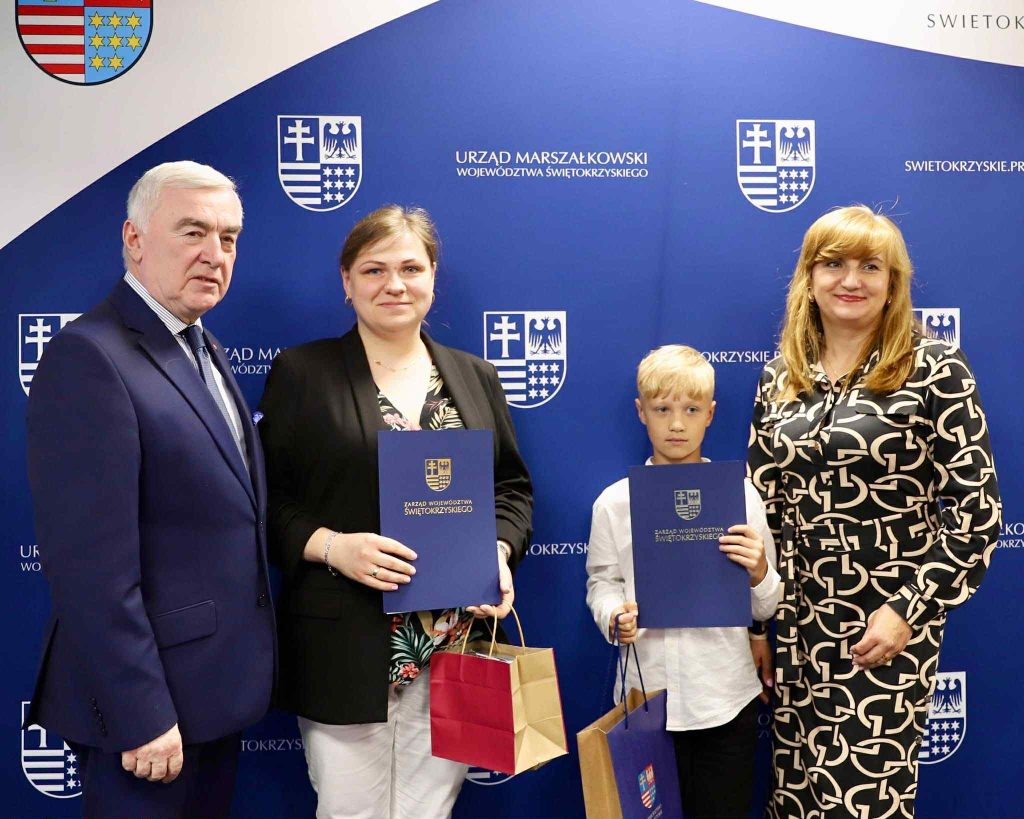 Fotografia przedstawia przewodniczącego sejmiku świętokrzyskiego oraz dwie kobiety i wyróżnionego chłopca na tle ścianki urzędu wojewódzkiegi województwa świętokrzyskiego, chłopiec i jedna kobieta trzymają granatowe teczki na dyplomy skierowane w stronę obiektywu, mają również torebki papierowe z prezentami