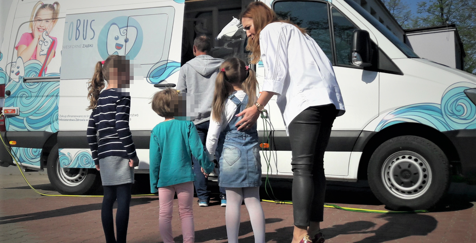 Grupa dzieci i dorosłych zbiera się wokół kolorowo udekorowanego samochodu stomatologicznego, biorąc udział w wydarzeniu promującym zdrowie społeczności.