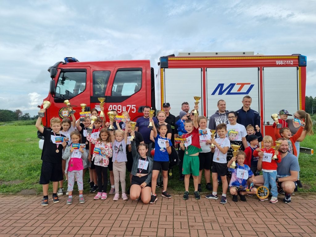 Grupa dzieci z dumą prezentuje swoje trofea i medale przed czerwonym wozem strażackim, świętując udane wydarzenie.