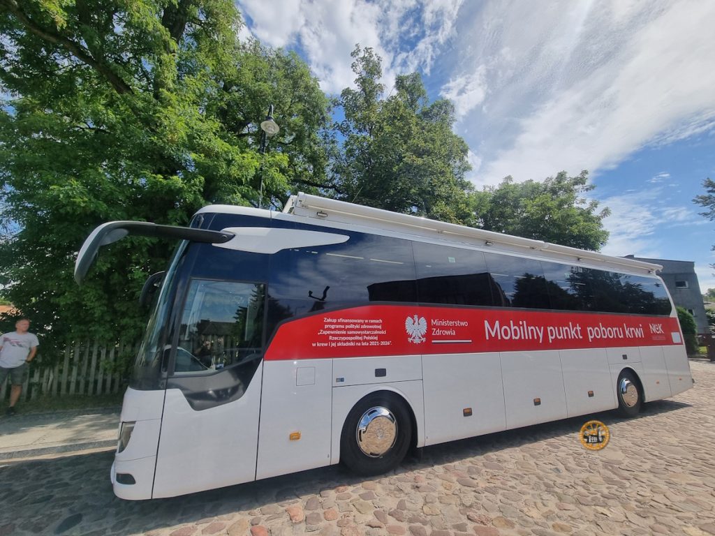 Mobilny autobus do pobierania krwi zaparkowany na brukowanej ulicy, ozdobiony czerwono-białym motywem i logo polskiego Ministerstwa Zdrowia.