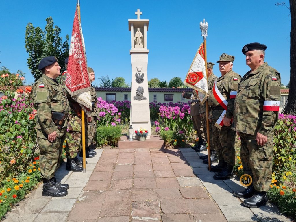 Żołnierze w strojach wojskowych stoją uroczyście obok pomnika udekorowanego kwiatami, trzymając flagi w spokojnym otoczeniu na świeżym powietrzu.