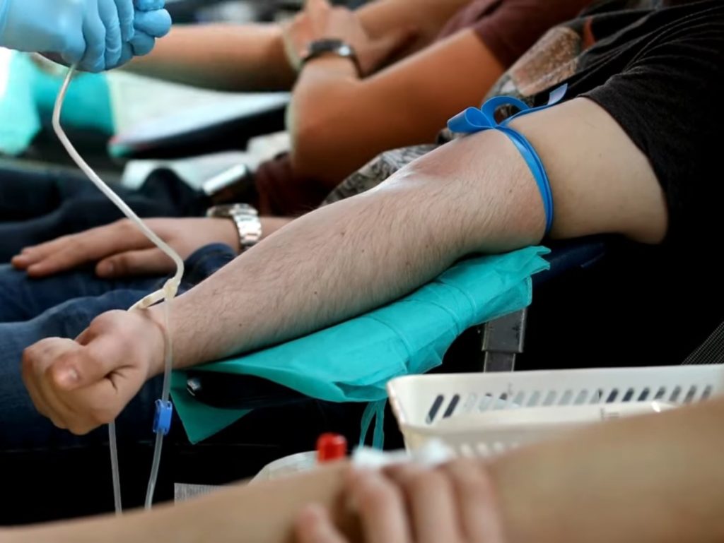 Zbliżenie na rękę osoby z niebieską opaską uciskową, przygotowującej się do oddania krwi, w towarzystwie innych dawców w placówce medycznej.