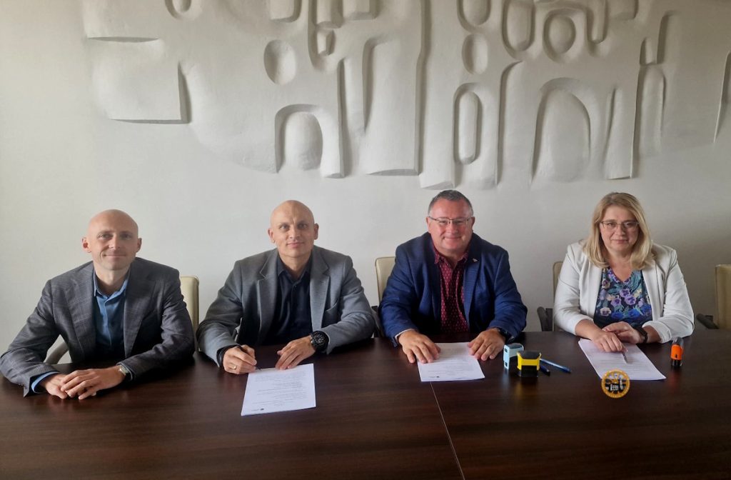 Cztery osoby siedzą przy stole konferencyjnym, przed nimi leżą dokumenty, a wokół nich znajduje się ściana o fakturowanej białej powierzchni.