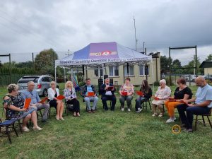 Grupa dorosłych siedzi w półokręgu, biorąc udział w spotkaniu na świeżym powietrzu, na tle zachmurzonego nieba i zieleni.