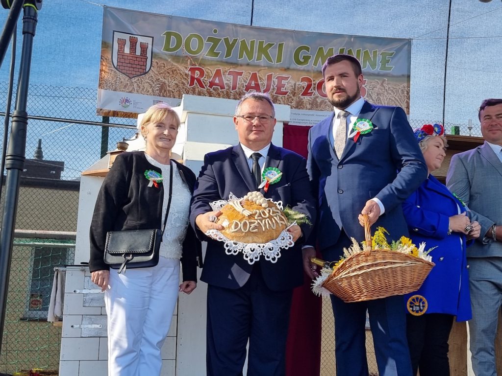 Grupa osób stoi na scenie podczas Dożynek Gminnych, trzymając ozdobiony bochenek chleba i ozdobny koszyk.