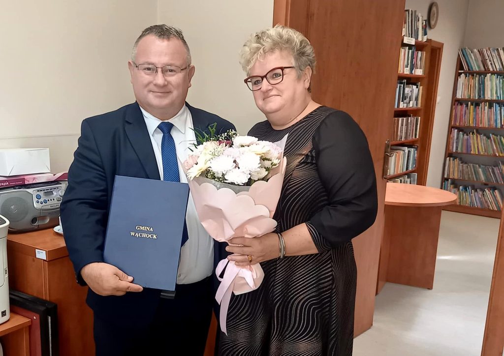 Mężczyzna w garniturze trzyma certyfikat, a kobieta w czarnej sukience trzyma bukiet kwiatów, świętując w ten sposób szczególną okazję, osoby są w pomieszczeniu, z tyłu na regałach znajdują się książki.