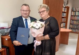 Mężczyzna w garniturze trzyma certyfikat, a kobieta w czarnej sukience trzyma bukiet kwiatów, świętując w ten sposób szczególną okazję, osoby są w pomieszczeniu, z tyłu na regałach znajdują się książki.