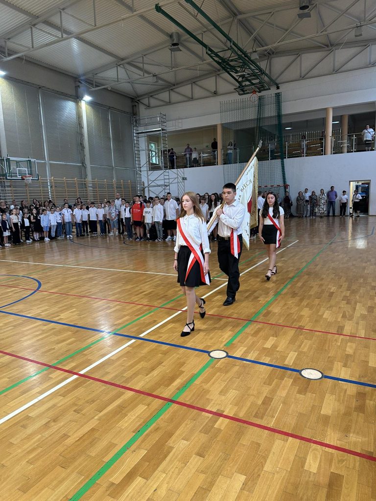 Uczniowie ubrani w formalne stroje niosą flagę podczas ceremonii w sali gimnastycznej, a uroczystość obserwuje grupa kolegów z klasy.
