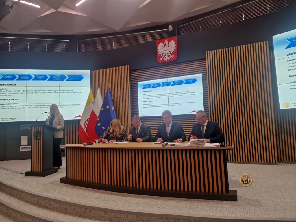 Sala konferencyjna, w której urzędnicy podpisują dokumenty, otoczona flagami Polski i Unii Europejskiej oraz ekranami wyświetlającymi informacje.