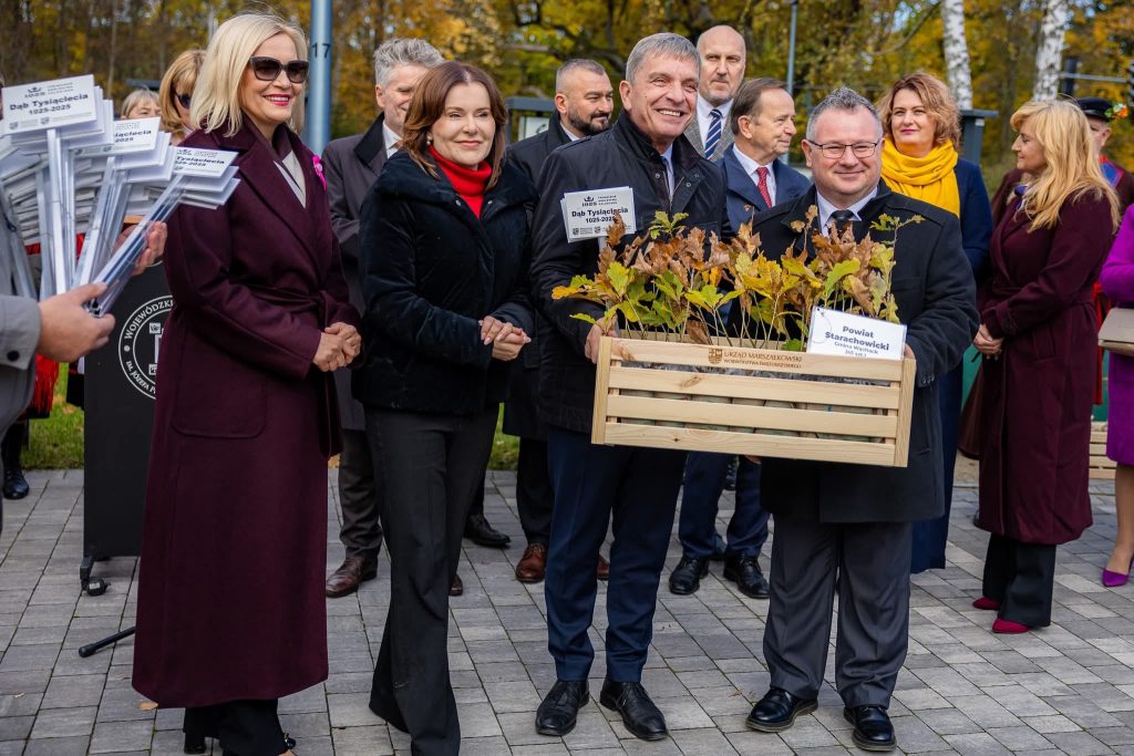 Grupa osób na imprezie plenerowej trzyma drewnianą skrzynię z sadzonkami oznaczonymi jako przeznaczone do sadzenia.