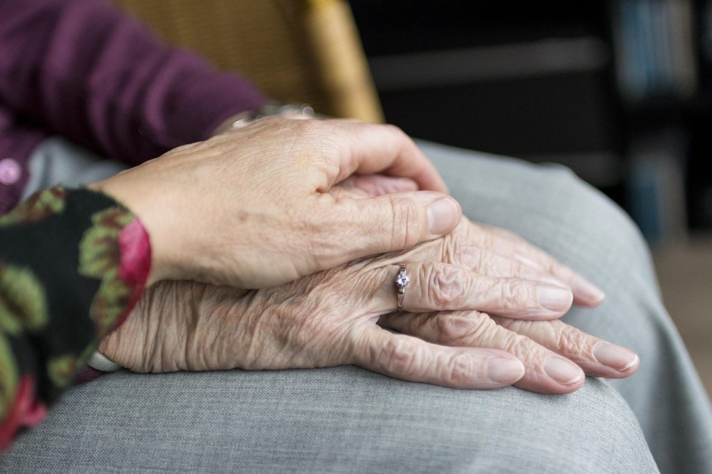 Dwie delikatnie splecione dłonie, odsłaniające starą skórę i pierścionek, symbolizujący więź i opiekę między pokoleniami.