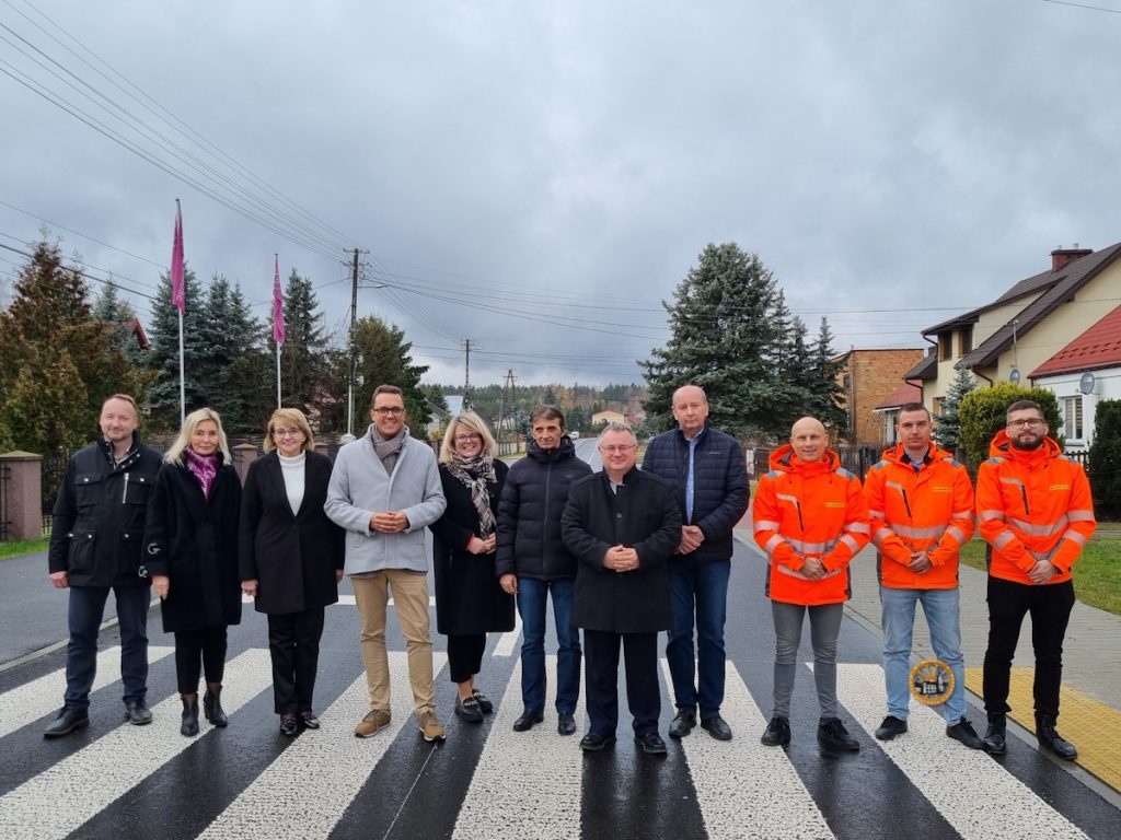 Zróżnicowana grupa osób stoi razem na przejściu dla pieszych, w tle widać budynki mieszkalne i drzewa, a pod zachmurzonym niebem.