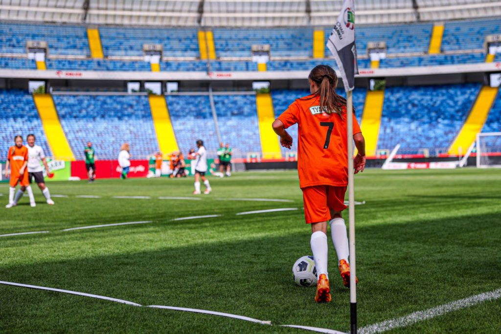 Młoda piłkarka w pomarańczowej koszulce przygotowuje się do wykonania rzutu rożnego na zielonym boisku podczas meczu na stadionie.