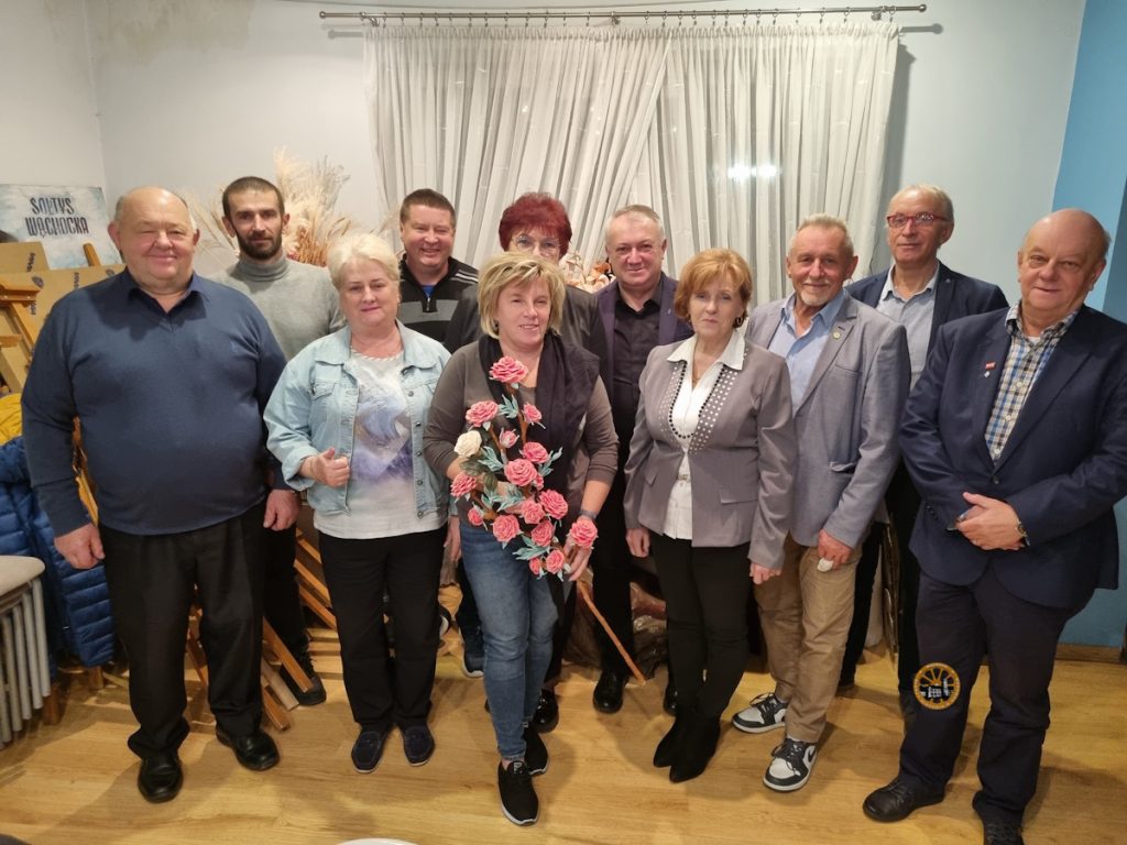 Grupa osób ubranych w różne style, pozujących razem w pomieszczeniu, w którym jedna osoba trzyma kompozycję kwiatową.