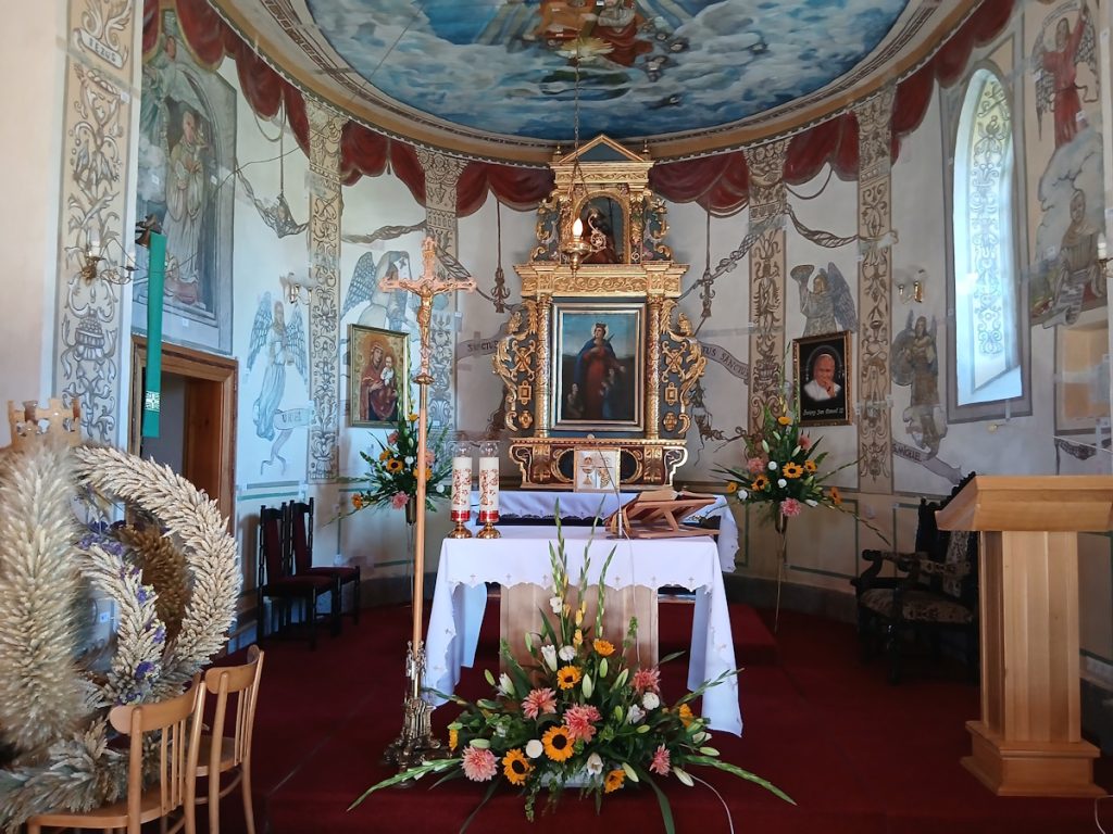Wnętrze kościoła z ozdobnym ołtarzem, kompozycjami kwiatowymi i malowidłami religijnymi na ścianach, tworzące spokojną atmosferę.
