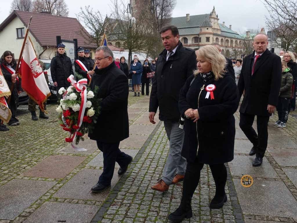 Grupa osób maszeruje razem, niektórzy trzymają flagi i wieniec kwiatowy, podczas publicznej uroczystości upamiętniającej na świeżym powietrzu.