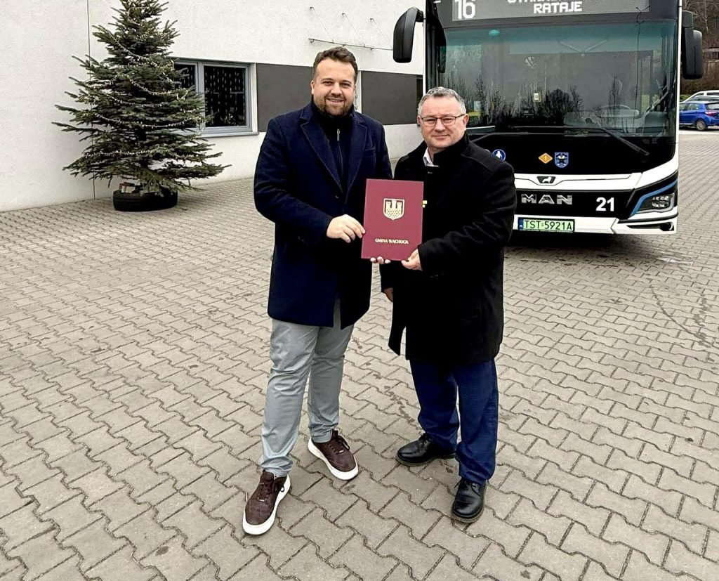 Dwóch mężczyzn stoi przed budynkiem urzędu miejskiego, trzymając dokument. W tle widać autobus z napisem „linia 16”.