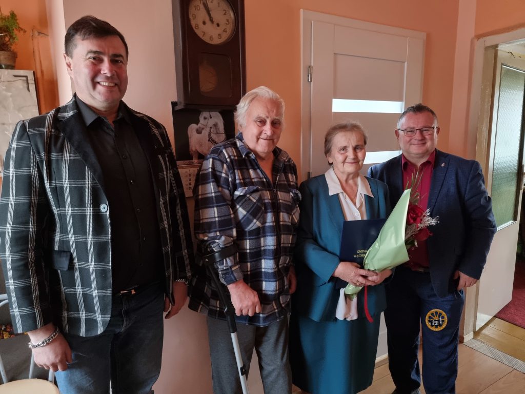 Kobieta trzyma bukiet i certyfikat, otoczona przez trzech mężczyzn, w przytulnym wnętrzu, prezentując moment świętowania.