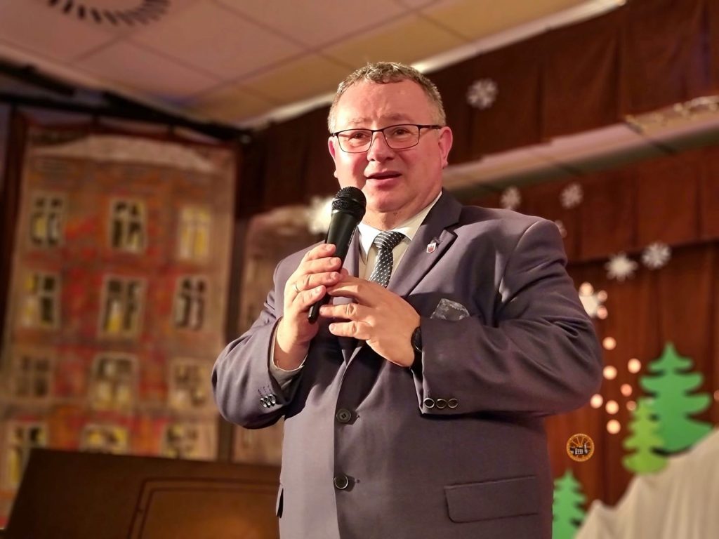 Osoba w garniturze trzyma mikrofon i przemawia na scenie udekorowanej świątecznymi elementami oraz na pomalowanym tle.