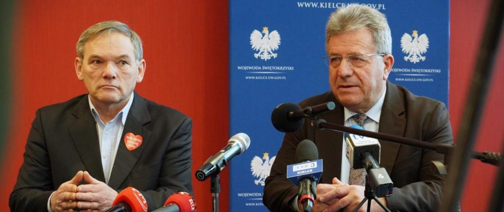 Dwóch mężczyzn siedzi przy stole z mikrofonami i kamerami na konferencji prasowej, na tle czerwonego tła i niebieskiego oficjalnego baneru.