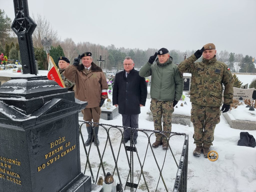 Cztery osoby w mundurach i płaszczach wojskowych salutują na ośnieżonym cmentarzu, w tle widoczne są nagrobki i pomniki.