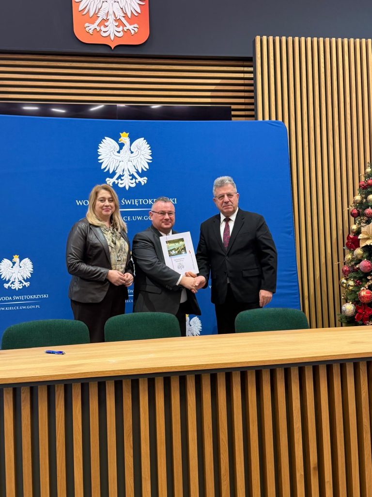 Formalne spotkanie, podczas którego trzy osoby ściskają sobie dłonie, trzymając w rękach certyfikat, na tle niebieskiego tła z polskim orłem.