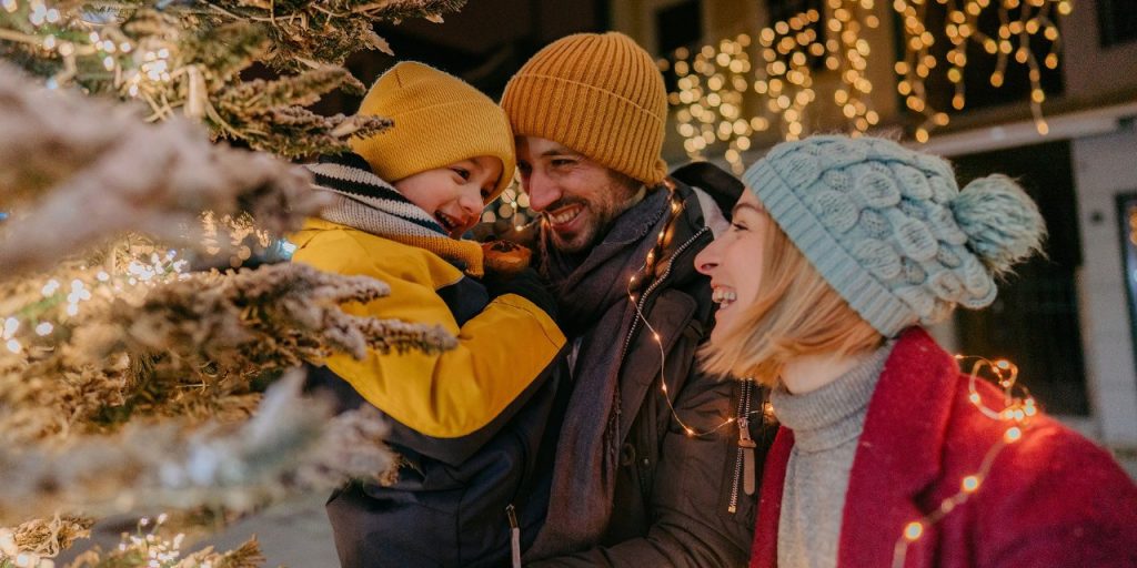 Rodzina zbiera się wokół udekorowanej choinki, nosi zimowe czapki, a delikatne światełka tworzą ciepłą, świąteczną atmosferę.