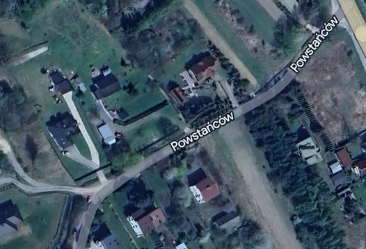 Zdjęcie satelitarne przedstawiające fragment ulicy Powstańców w Wąchocku wraz z zabudową jednorodzinną, przydomowymi ogrodami oraz terenami zielonymi wzdłuż drogi.