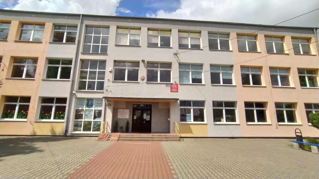 Budynek szkoły o trzech kondygnacjach, z jasną elewacją w odcieniach beżu i szarości. Fasada jest symetryczna, z równomiernie rozmieszczonymi, dużymi prostokątnymi oknami w białych ramach.

W centralnej części znajduje się główne wejście z niewielkim zadaszeniem, do którego prowadzą schody oraz podjazd z żółtymi poręczami. Nad drzwiami umieszczona jest czerwona tablica informacyjna.

Przed budynkiem widoczny jest szeroki, wybrukowany plac z kostki brukowej. Na parterze, w oknach po lewej stronie, znajdują się kolorowe dekoracje. Nad budynkiem częściowo widoczne jest błękitne niebo z białymi chmurami.