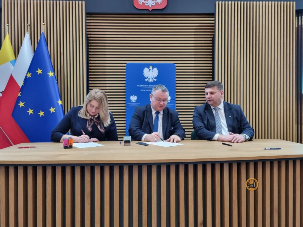 Trzy osoby podpisujące dokumenty przy drewnianym stole w sali urzędowej; w tle godło Polski, flaga Polski i Unii Europejskiej oraz tablica Wojewody Świętokrzyskiego.