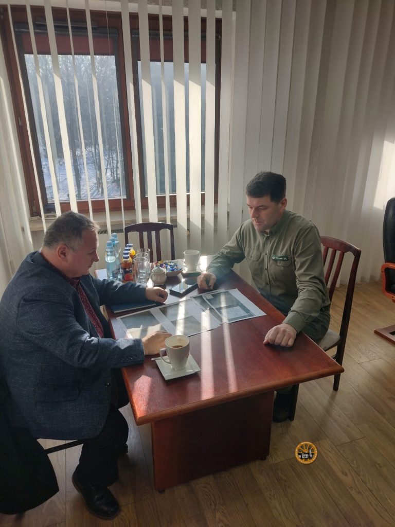 Dwóch mężczyzn siedzi przy drewnianym stole w jasnym biurze i analizuje rozłożone na blacie dokumenty oraz wydruki planów. Na stole stoją filiżanki z kawą, butelki z wodą i notatnik, a za nimi widać duże okno z pionowymi żaluzjami i zimowy krajobraz za szybą.