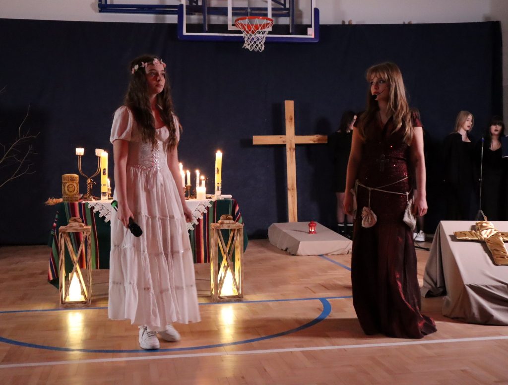 Scena teatralna w sali gimnastycznej. Dwie młode kobiety w stylizowanych, długich sukniach stoją naprzeciw siebie, jedna w jasnej, druga w ciemnej kreacji. Między nimi ustawiono stół ze świecami i drewniany krzyż, a w tle widoczne są kolejne osoby oraz elementy scenografii tworzące nastrój obrzędu