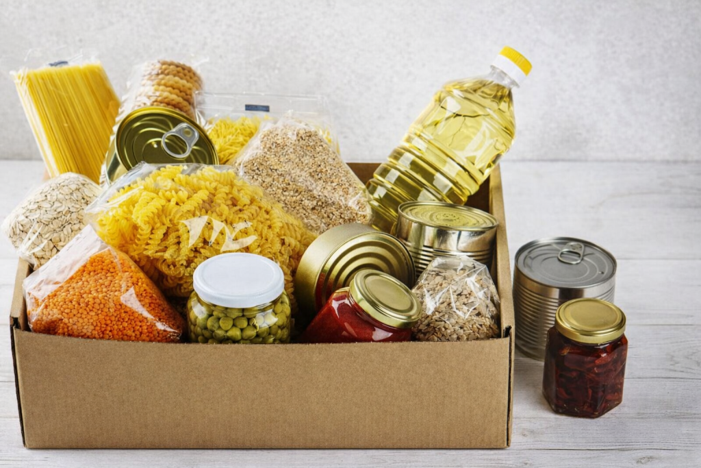 Kartonowe pudełko wypełnione produktami spożywczymi o długim terminie przydatności, ustawione na jasnym tle w poziomym kadrze. W środku widoczne są opakowania makaronu spaghetti i świderków, czerwona soczewica w przezroczystym woreczku, płatki owsiane, kasza oraz kilka metalowych puszek. Z boku znajduje się butelka oleju roślinnego, słoik zielonego groszku i słoik z czerwonymi warzywami w zalewie. Całość oświetlona miękkim, naturalnym światłem.
