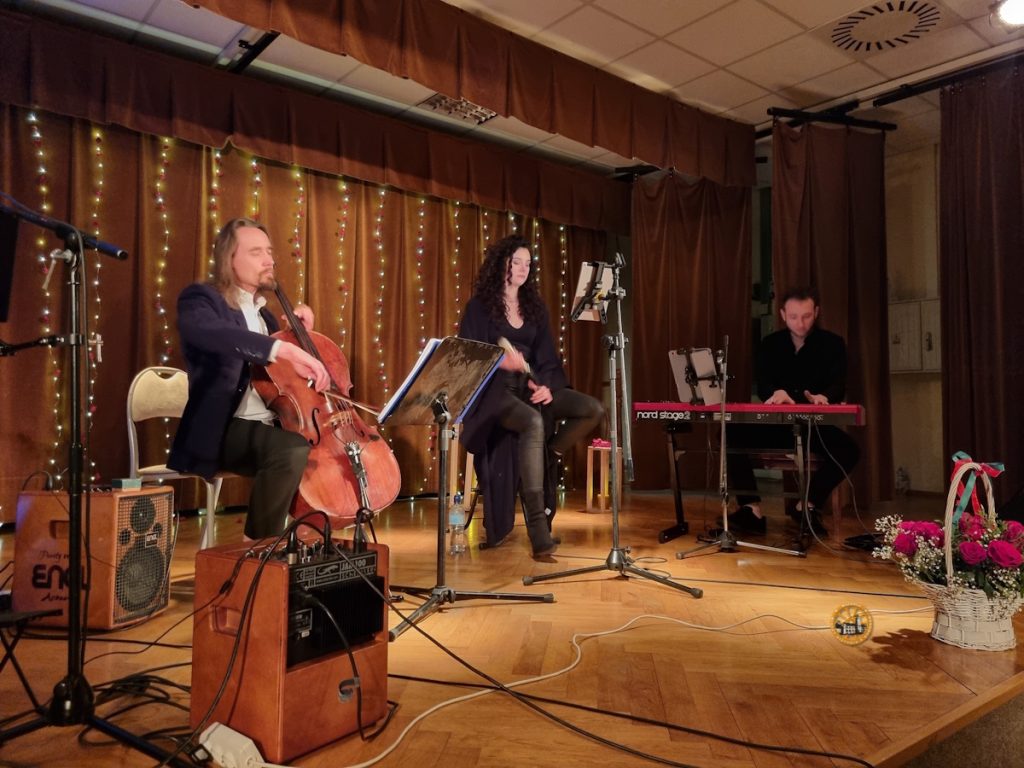 Na niewielkiej scenie podczas kameralnego koncertu siedzi troje muzyków. Po lewej wiolonczelista gra na instrumencie, w centrum wokalistka siedzi przy mikrofonie i pulpicie z nutami, a po prawej pianista gra na klawiaturze Nord Stage. W tle wiszą brązowe kotary ozdobione pionowymi girlandami światełek. Na scenie stoją statywy mikrofonowe, wzmacniacze, przewody oraz kosz z kwiatami przy krawędzi sceny.