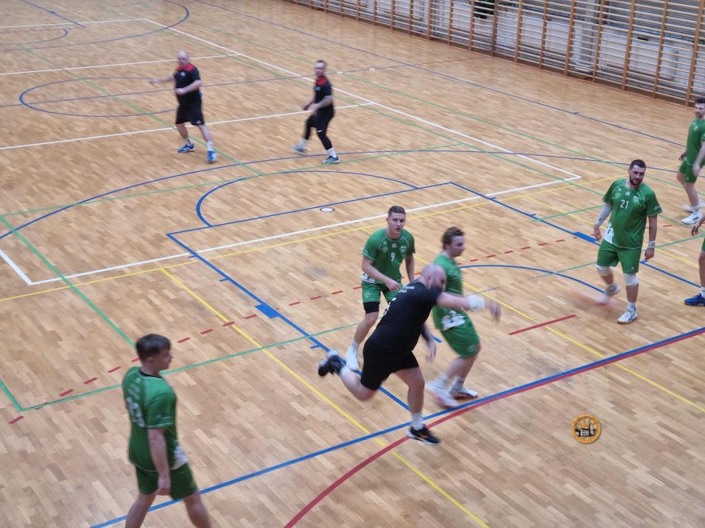 Zawodnicy dwóch drużyn rozgrywają akcję podczas meczu piłki ręcznej w hali sportowej. Gracz w czarnym stroju wykonuje rzut w wyskoku, otoczony przez zawodników w zielonych koszulkach. Mecz odbywa się na drewnianym parkiecie z wyraźnie zaznaczonymi liniami boiska