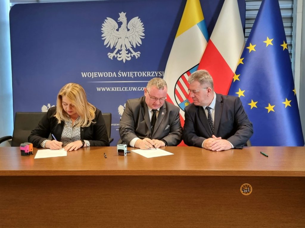 Trzy osoby w formalnych strojach siedzą przy drewnianym stole i podpisują dokumenty. W tle znajduje się niebieska ścianka z godłem Polski i napisem „Wojewoda Świętokrzyski”, a obok ustawione są flagi Polski, województwa świętokrzyskiego oraz Unii Europejskiej. Sytuacja wygląda na oficjalne podpisanie dokumentów w urzędzie.