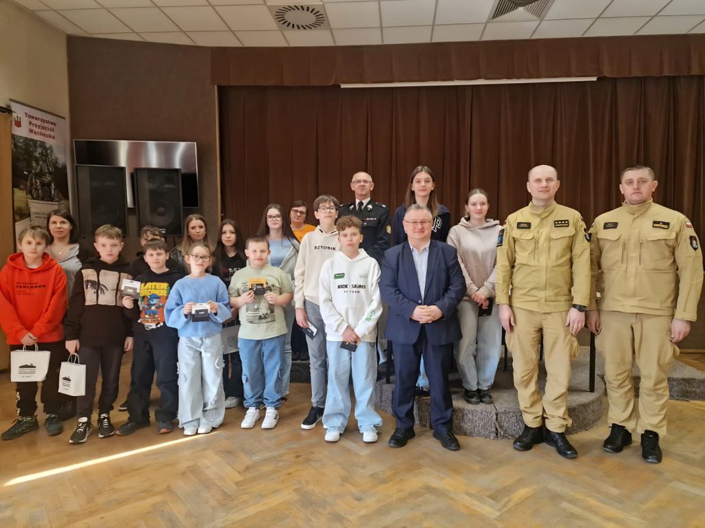 Grupa dzieci, młodzieży oraz dorosłych stoi razem na niewielkiej scenie w sali z brązowymi zasłonami. Na środku widoczny mężczyzna w granatowym garniturze, obok niego osoby w mundurach służb ratowniczych. Uczestnicy trzymają dyplomy lub upominki. W tle znajdują się głośniki oraz baner Towarzystwa Przyjaciół Wąchocka. Zdjęcie wykonane podczas oficjalnego spotkania lub wydarzenia edukacyjnego.