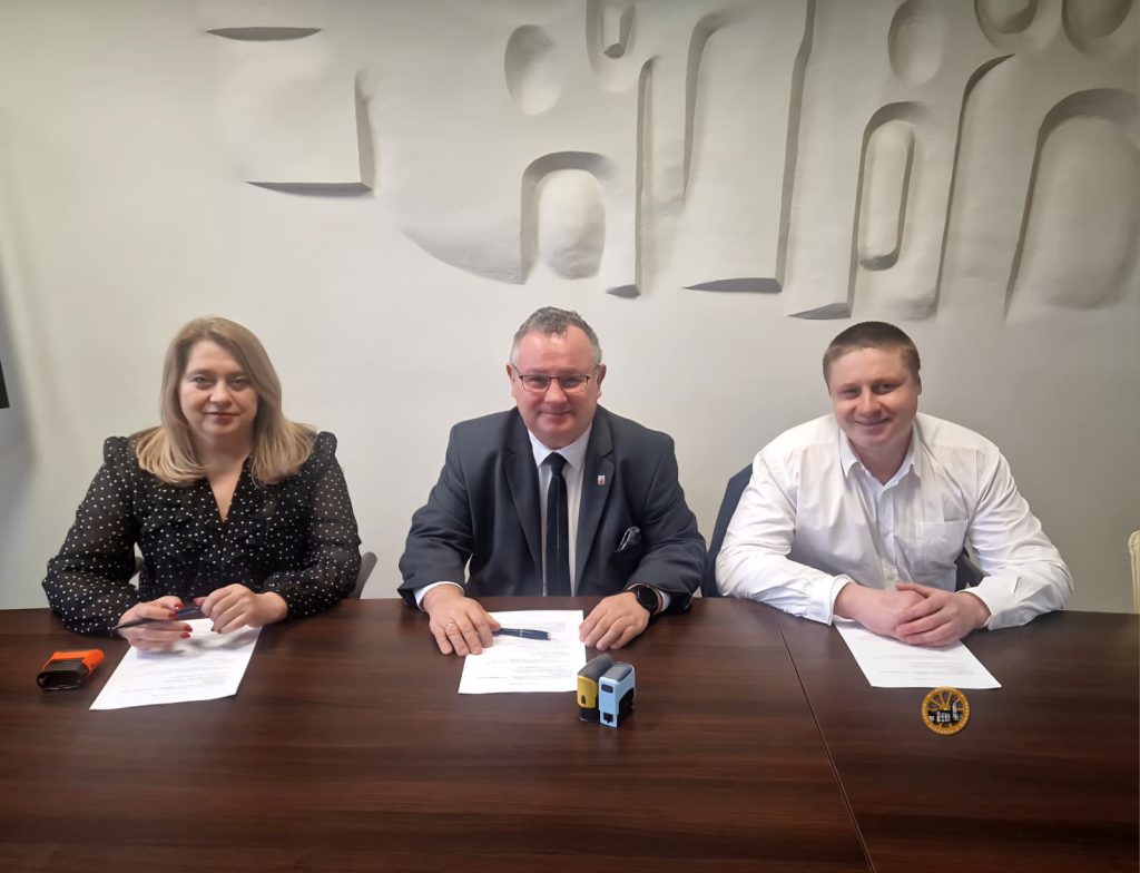 Trzy osoby siedzą przy drewnianym stole podczas oficjalnego spotkania lub podpisywania dokumentów. W centrum znajduje się mężczyzna w garniturze i krawacie, po jego bokach siedzą kobieta w czarnej bluzce w kropki oraz mężczyzna w białej koszuli. Na stole leżą dokumenty, długopisy i pieczątki, a w tle widoczna jest jasna ściana z dekoracyjnym reliefem